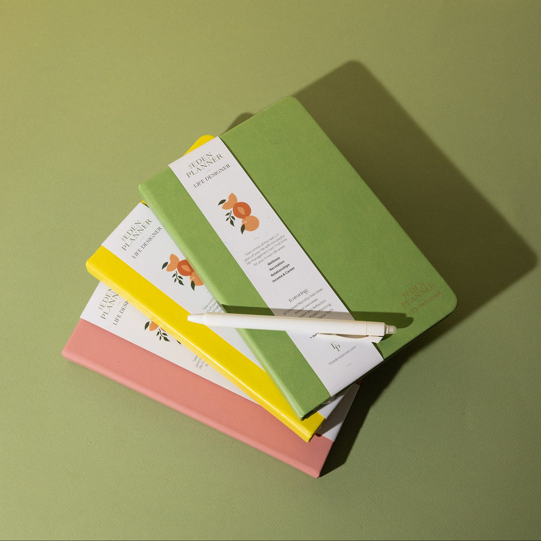 Three hardcover planners stacked on top of each other in sage green, yellow, and dusty pink, with a white pen resting on the top notebook. Each notebook has a white paper band with orange peach illustrations and the brand name 'Eden Planner Life Designer'.